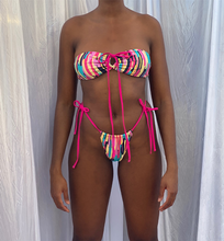 Load image into Gallery viewer, model wearing the paint palate bikini bandeau style on the bust, paired with high thigh thong multi colour bottoms with bright pink hip straps. 
