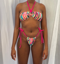 Load image into Gallery viewer, model wearing the paint palate bikini bandeau style on the bust, paired with high thigh thong multi colour bottoms with bright pink hip straps. 
