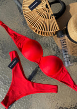 Load image into Gallery viewer, A red bikini set with no straps, placed on a flat surface alongside a straw hat and a water bottle, with a visible brand tag on the bottom left.
