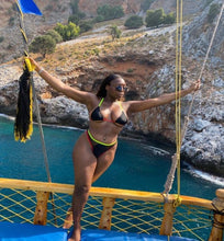 Load image into Gallery viewer, A woman standing on a boat wearing a black and red bikini with yellow accents, posing with one hand extended and a scenic ocean background.
