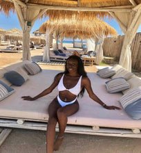 Load image into Gallery viewer, A woman sitting on a sun lounger under a thatched umbrella, wearing a white one-piece swimsuit.
